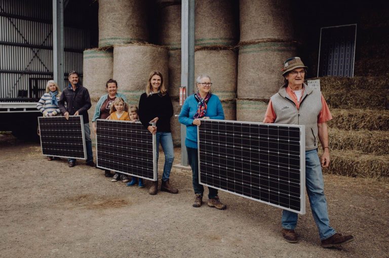 6 adults and two children carry solar panels with a hayshed in the background.