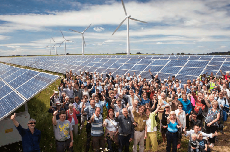 A large group of people stand happily in front of a solar farm with wind turbine in the background